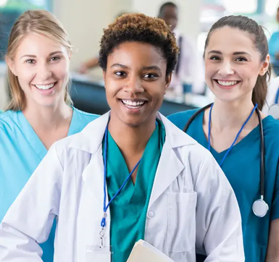 Three female nurses in scrubs standing together in a hospital corridor, smiling and engaged in conversation.